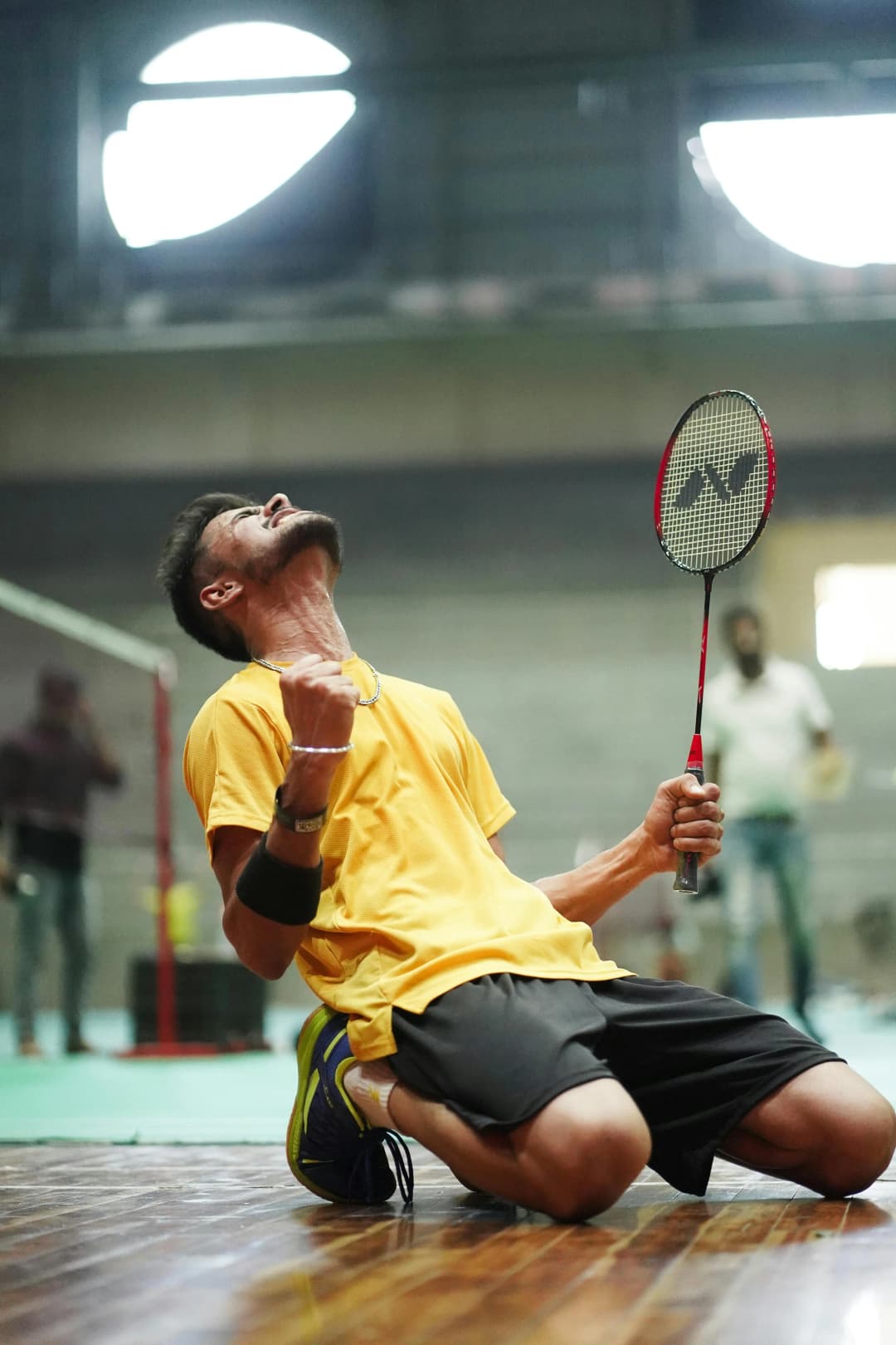 Joueur de badminton à genoux, raquette à la main, exprimant une intense émotion sur un terrain en salle.