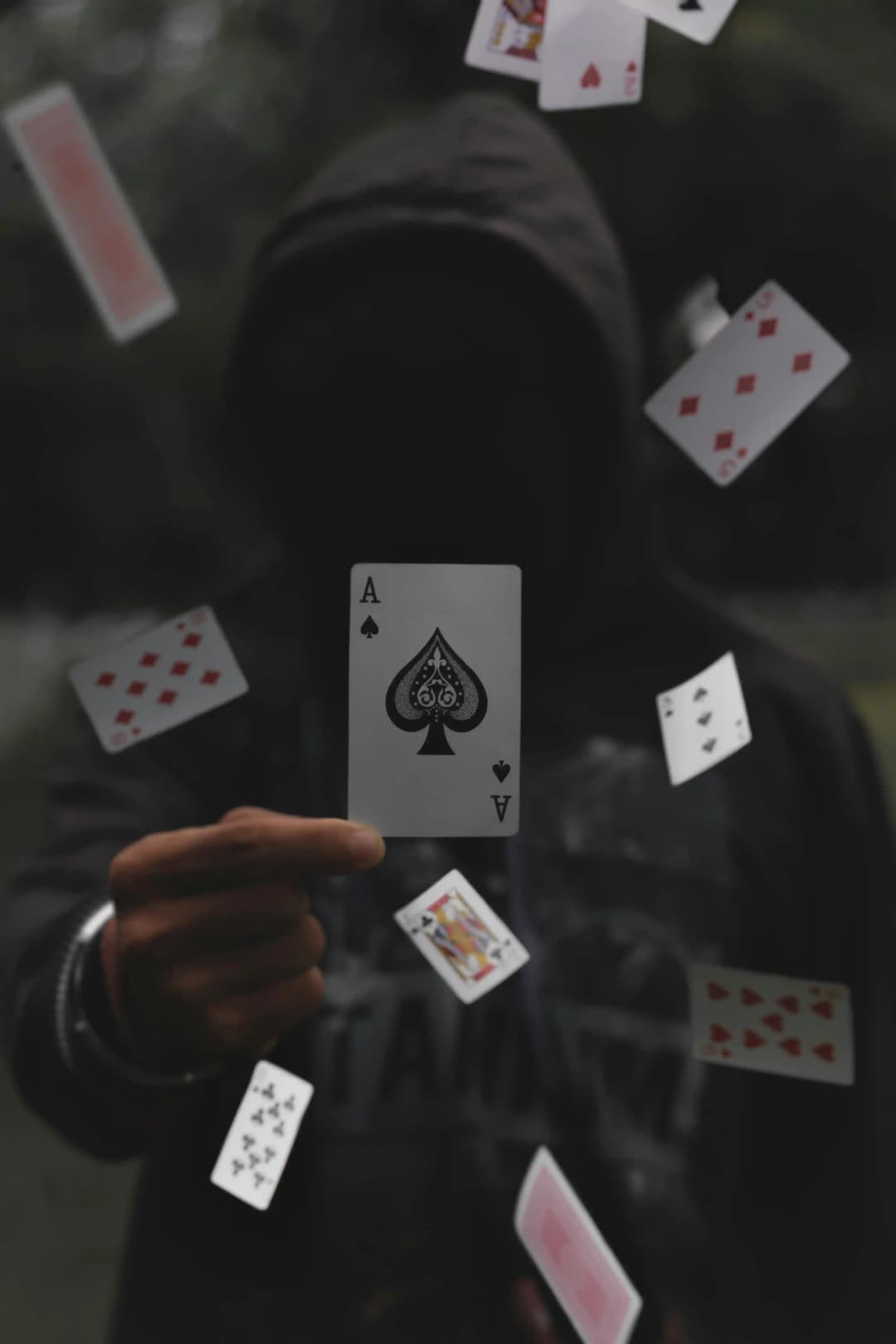 Personne encapuchonnée tenant une carte As de pique, entourée de cartes flottant dans les airs.